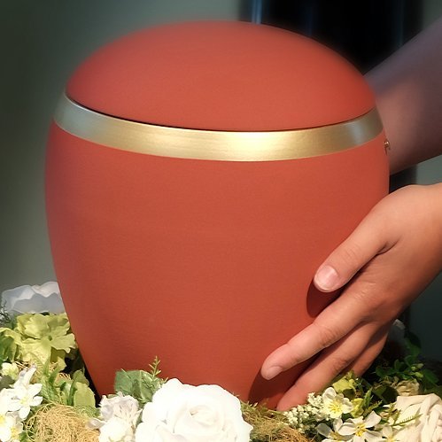 Rote Urne auf Blumenkranz von Händen umfasst